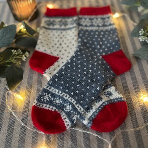 May include: A pair of knitted wool socks with a red, white and blue snowflake pattern. The socks are folded and displayed on a striped fabric background.