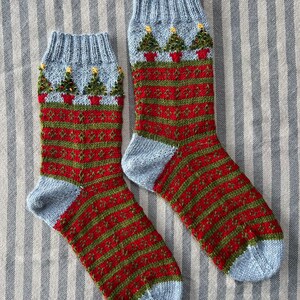 Peut inclure: Une paire de chaussettes de No&euml;l tricot&eacute;es. Les chaussettes ont un bord et un bout du pied bleu clair, avec un motif &agrave; rayures rouges et vertes sur le corps. Le bord pr&eacute;sente de petits arbres de No&euml;l en pots avec des accents d'&eacute;toiles jaunes.
