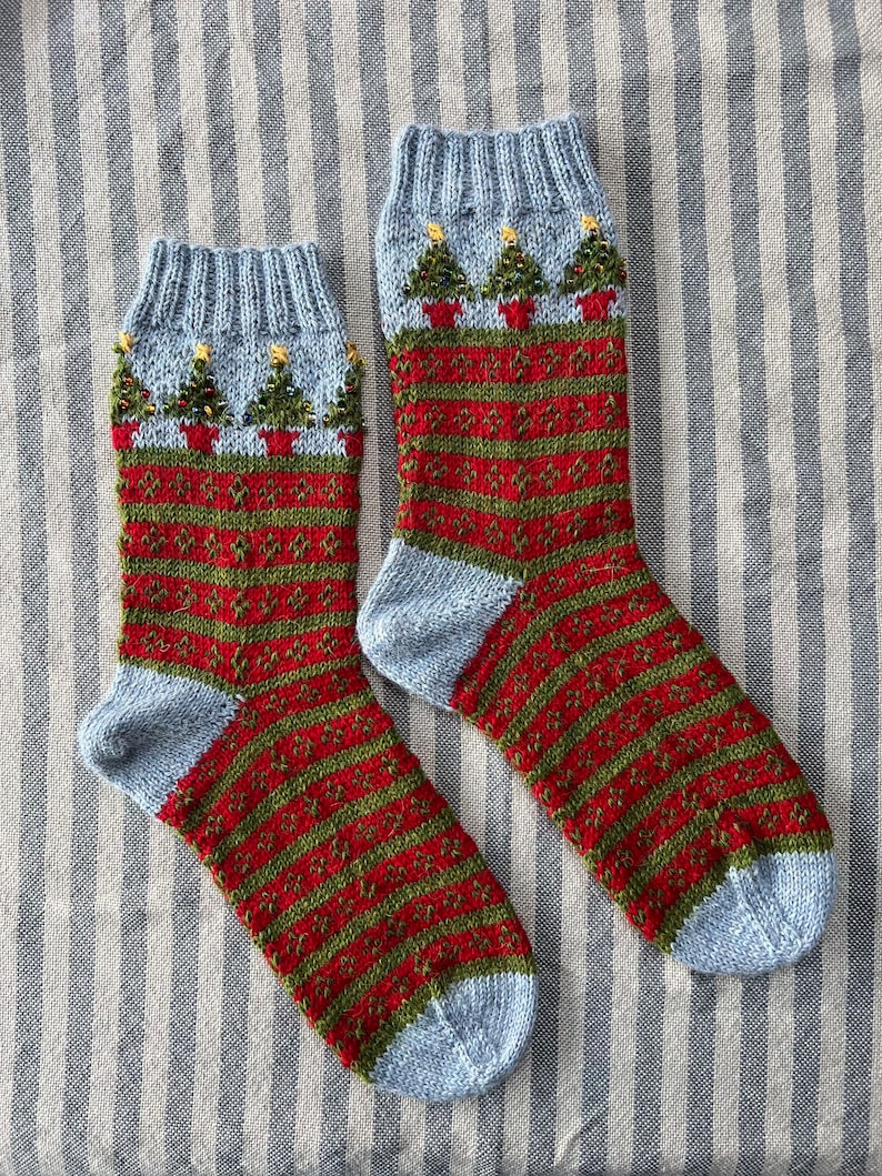 Peut inclure: Une paire de chaussettes de No&euml;l tricot&eacute;es. Les chaussettes ont un bord et un bout du pied bleu clair, avec un motif &agrave; rayures rouges et vertes sur le corps. Le bord pr&eacute;sente de petits arbres de No&euml;l en pots avec des accents d'&eacute;toiles jaunes.