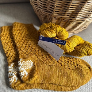 May include: Mustard yellow knitted socks with a white snowflake design on the ankle. A ball of matching yarn and a tag that reads "013 ORO ULTIMATE SOCK" are next to the socks. A wicker basket is in the background.