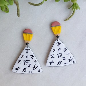 May include: A pair of white triangle earrings with a yellow and pink pencil topper. The triangles are covered in mathematical symbols such as pi, square root, summation, and infinity.