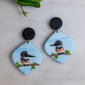 May include: A pair of handmade earrings featuring a bird design. Each earring has a light blue, diamond-shaped pendant with a black bird perched on a branch. The bird has black, white, and orange details. The earrings have black circular tops.