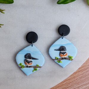 May include: A pair of handmade earrings featuring a bird design. Each earring has a black circular top and a light blue, diamond-shaped pendant. The pendant showcases a detailed bird perched on a branch, with a black, white, and orange color scheme.