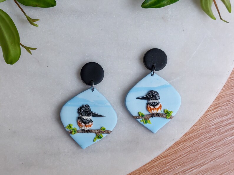 May include: A pair of handmade earrings featuring a bird design. Each earring has a black circular top and a light blue, diamond-shaped pendant. The pendant showcases a detailed bird perched on a branch, with a black, white, and orange color scheme.