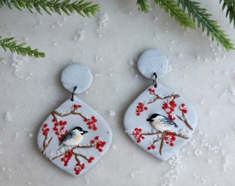 May include: A pair of dangle earrings featuring a light gray diamond-shaped base with a hand-painted scene of a small black and white bird perched on a brown branch with bright red berries and white snow accents, attached to a circular gray stud.
