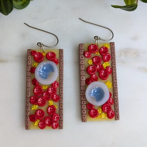 May include: A pair of earrings featuring a white background with red and yellow circles and a blue circle in the centre. The earrings are made of polymer clay and have silver hooks.