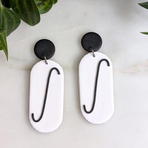 May include: A pair of white dangle earrings with black accents. Each earring features a black circle at the top and a white oval with a black curved line in the shape of an integral symbol.