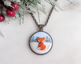 May include: A round pendant necklace with a winter scene featuring an orange fox in the snow. The pendant has a silver-colored frame and a dark chain. The background includes a blue sky and green trees. Red berries and green branches are in the upper left corner.