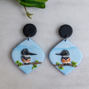 May include: A pair of handmade earrings featuring a bird design. Each earring has a black circular top and a light blue, diamond-shaped pendant. The pendant showcases a detailed bird perched on a branch, with green leaves and a blue sky background.