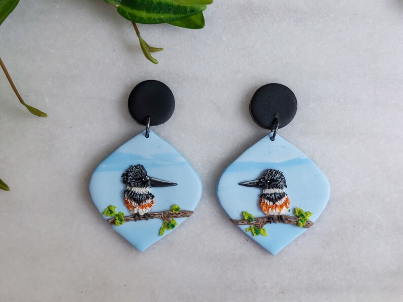 May include: A pair of handmade earrings featuring a bird design. Each earring has a black circular top and a light blue, diamond-shaped pendant. The pendant showcases a detailed bird perched on a branch, with green leaves and a blue sky background.