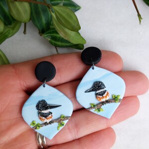 May include: A pair of handmade earrings featuring a bird design. Each earring has a light blue, diamond-shaped pendant with a black and white bird perched on a branch. The earrings have black circular studs.