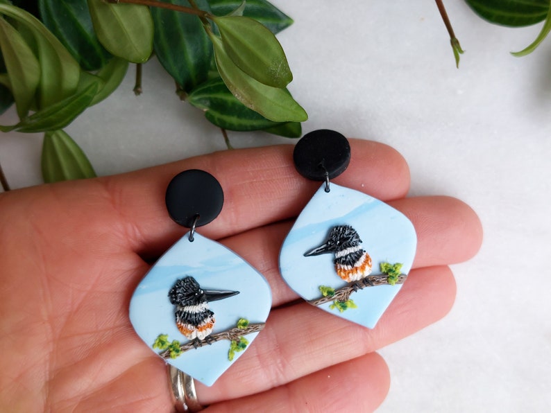 May include: A pair of handmade earrings featuring a bird design. Each earring has a light blue, diamond-shaped pendant with a black and white bird perched on a branch. The earrings have black circular studs.