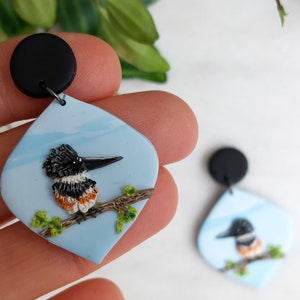 May include: A pair of handcrafted earrings featuring a bird design. Each earring has a black top and a light blue, teardrop-shaped pendant. The pendant showcases a detailed bird perched on a branch with green leaves.