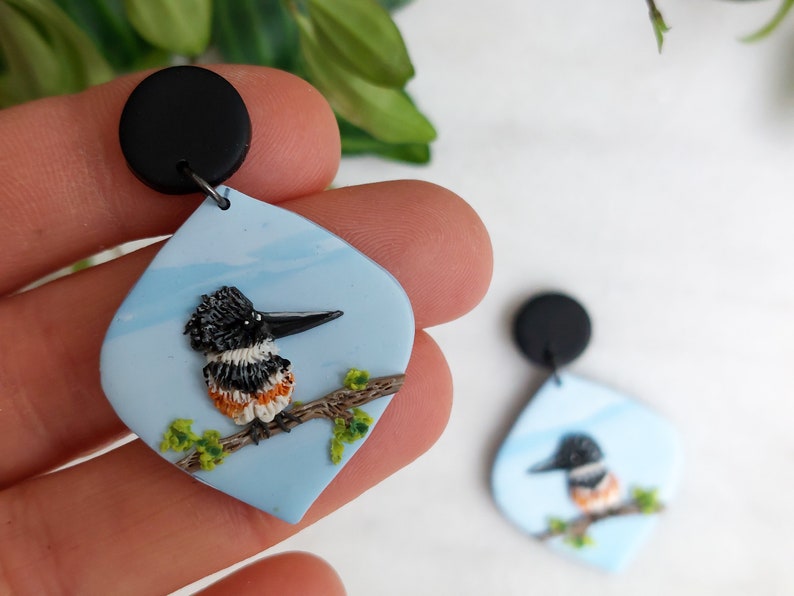 May include: A pair of handcrafted earrings featuring a bird design. Each earring has a black top and a light blue, teardrop-shaped pendant. The pendant showcases a detailed bird perched on a branch with green leaves.