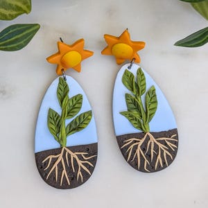 May include: A pair of handmade polymer clay earrings featuring a blue sky, green plant with roots, and a yellow sun. The earrings are oval shaped.