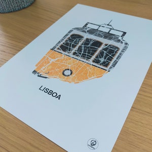 May include: A white print with a yellow and black tram design. The tram is made up of the city map of Lisbon, Portugal. The print has the word "LISBOA" and a small logo at the bottom.