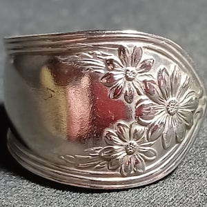 May include: A silver ring made from a repurposed spoon with a floral design. The ring features three stylized flowers with a raised design.
