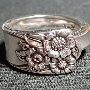May include: A silver spoon ring with a floral design. The ring is made from a spoon with a floral pattern on the front. The spoon is bent into a ring shape.
