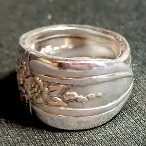 May include: A silver spoon ring with a floral design. The ring is made from a bent spoon and features a detailed floral pattern.