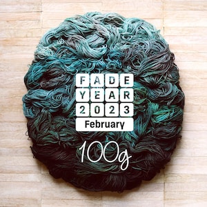 Puede incluir: Un gran ovillo circular de hilo teñido a mano en tonos de verde azulado, verde y negro. El hilo está dispuesto sobre una superficie de madera clara. Un gráfico cuadrado blanco muestra "FADE YEAR 2023 February" y "100g".