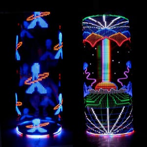 May include: Two illuminated cylindrical light fixtures. One features blue and orange cross-shaped designs, while the other displays a colorful rainbow-like pattern with red, green, and blue lights. The background is black.