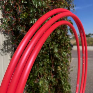 Puede incluir: Tres aros de hula rojos brillantes están dispuestos en arco, con un exuberante arbusto verde de fondo. Los aros son lisos y parecen estar hechos de un material flexible. La imagen está tomada al aire libre en un día soleado.