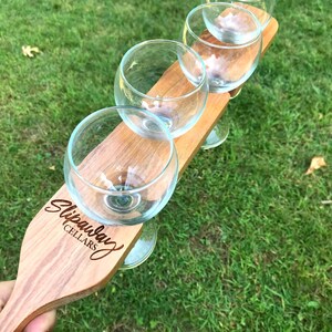 Puede incluir: Paleta de degustación de vino de madera con cuatro vasos pequeños unidos. La paleta está grabada con las palabras "Slip Away Cellars".