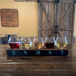May include: Five clear glass tasting glasses with different coloured liquids, numbered 5, 9, 10, 4, and 7 on a wooden board.
