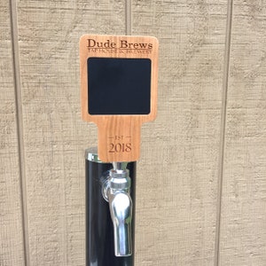 May include: Wooden tap handle with a chalkboard sign. The sign says "Dude Brews Tap House & Brewery - Est. 2018".