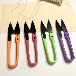 May include: Four pairs of small, colourful fabric scissors with black blades. The scissors are arranged in a row from left to right: orange, pink, green, and purple.