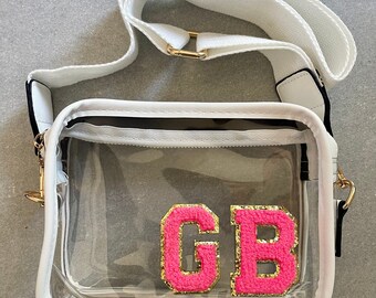 Puede incluir: Un bolso transparente de plástico con correa cruzada, correa blanca y ribete blanco. El bolso tiene cierre de cremallera y presenta dos letras de fieltro rosa con ribete de purpurina dorada que deletrean "GB".