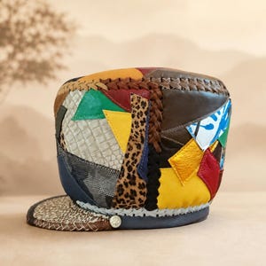 May include: A multi-coloured patchwork cap constructed from various leather textures and patterns, including leopard print, crocodile-embossed sections, and solid panels in yellow, green, red, blue, and brown, featuring braided leather trim details.