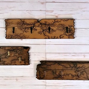 May include: Three wooden key racks with a burnt wood design and metal hooks. The racks are different sizes and are mounted on a white wood wall.
