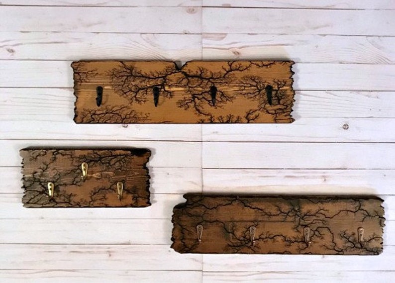 May include: Three wooden key racks with a burnt wood design and metal hooks. The racks are different sizes and are mounted on a white wood wall.