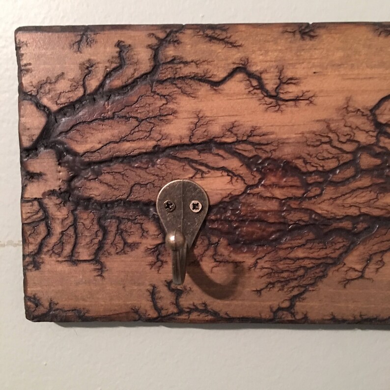 May include: A wooden wall hook with a dark brown wood grain and a black lightning bolt pattern. The hook is made of metal and has a dark bronze finish.