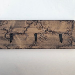 May include: A wooden key holder with a rustic, weathered finish and three black metal hooks. The wood has a unique crackled pattern.