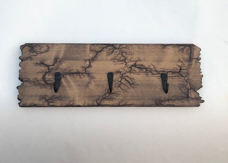 May include: A wooden key holder with a rustic, weathered finish and three black metal hooks. The wood has a unique crackled pattern.