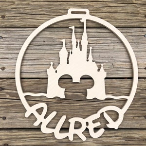 May include: A wooden ornament shaped like a circle with a silhouette of Cinderella's castle and Mickey Mouse ears inside. The word "ALLURED" is written in a whimsical font below the castle.