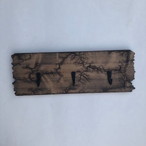 May include: A rustic wooden key holder with three black metal hooks. The wood has a dark brown stain and a unique crackled finish.