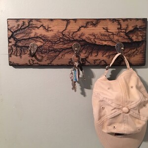 May include: A wooden key rack with a burnt wood design and three metal hooks. A set of keys and a beige baseball cap are hanging on the hooks.