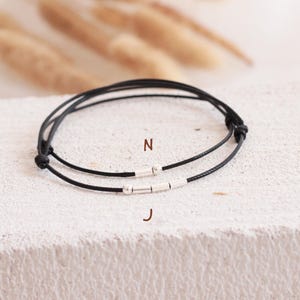 Peut inclure: Deux bracelets en cordon noir avec des perles cylindriques argentées. L'un des bracelets porte la lettre "N" et l'autre la lettre "J" sur le cordon. Les bracelets sont présentés sur une surface blanche texturée.