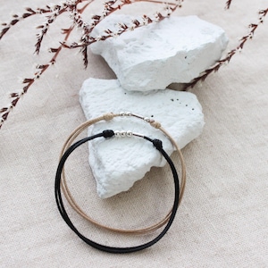 May include: Two minimalist bracelets, one black and one beige, with silver-toned metal accents. The bracelets are circular and rest on a white, textured surface. A sprig of dried flowers is in the background.