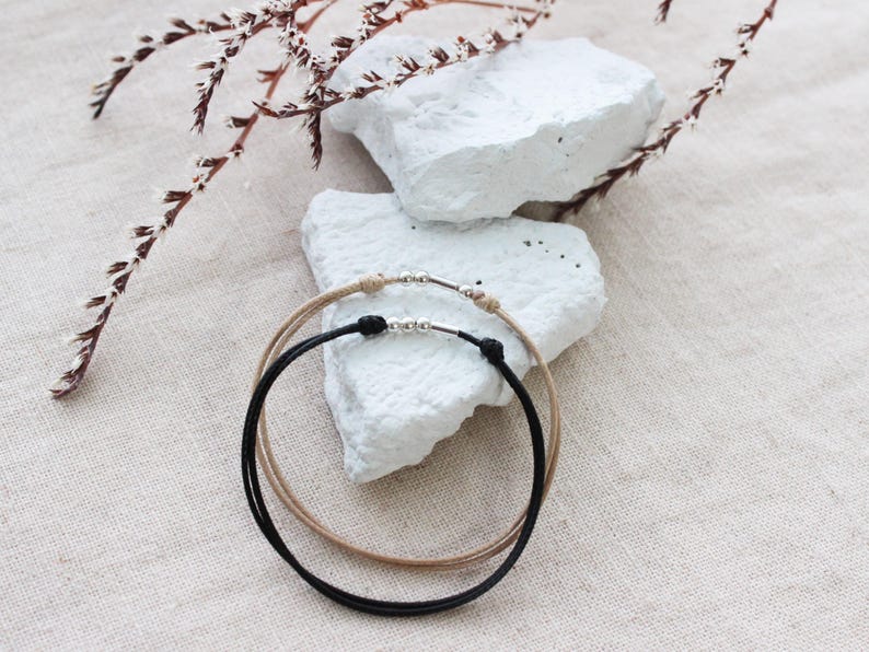 Peut inclure: Deux bracelets minimalistes, un noir et un beige, avec des accents m&eacute;talliques argent&eacute;s. Les bracelets sont circulaires et reposent sur une surface blanche textur&eacute;e. Une brindille de fleurs s&eacute;ch&eacute;es est en arri&egrave;re-plan.