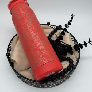 May include: A red insulated water bottle with a gold floral pattern. The bottle is in a black decorative basket with black beads and a tassel. The bottle has a red lid and base.