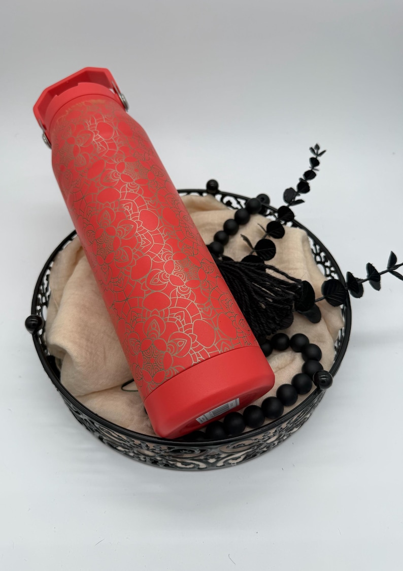 May include: A red insulated water bottle with a gold floral pattern. The bottle is in a black decorative basket with black beads and a tassel. The bottle has a red lid and base.