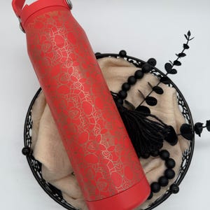 May include: A red insulated water bottle with a gold floral pattern. The bottle has a red handle and base. It rests in a black wire basket with a beige cloth, black beads, and decorative sprigs.