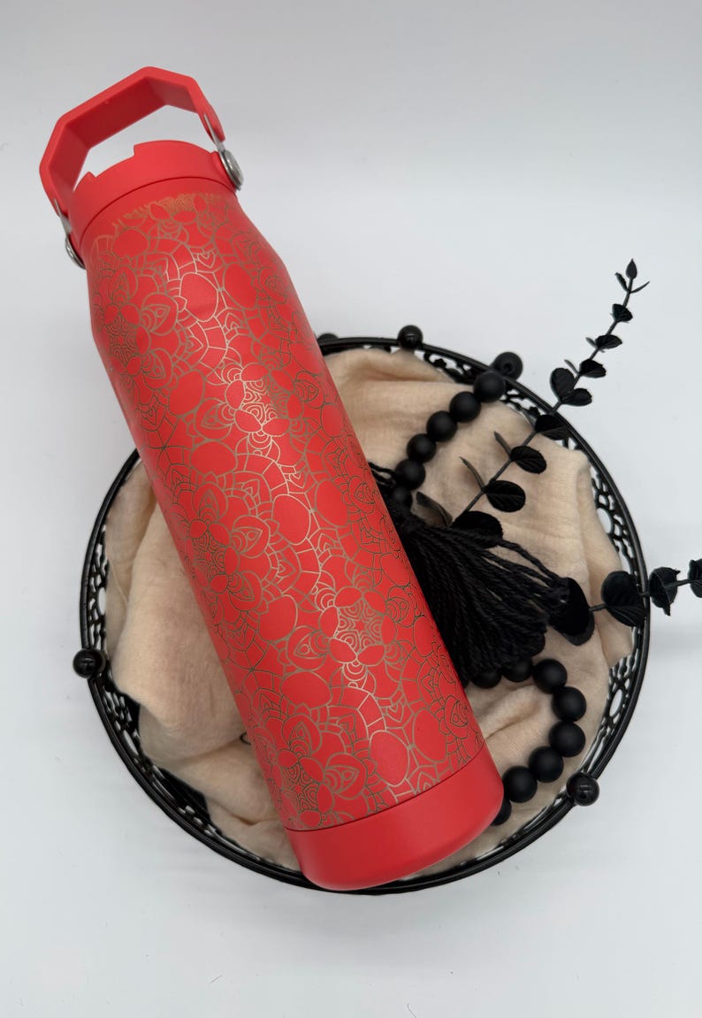 May include: A red insulated water bottle with a gold floral pattern. The bottle has a red handle and base. It rests in a black wire basket with a beige cloth, black beads, and decorative sprigs.