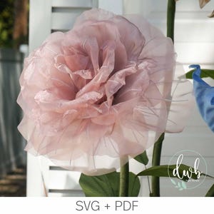 May include: A large, light pink paper flower with delicate, layered petals. The flower is mounted on a green stem with green leaves. The image also includes a blue and green flower. The text "SVG + PDF" is visible at the bottom.