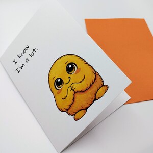 May include: A folded greeting card with a cartoon illustration of a yellow, fluffy creature with large eyes and a smiling expression. The text "I know I'm a lot" is printed on the card. An orange envelope is visible in the background.