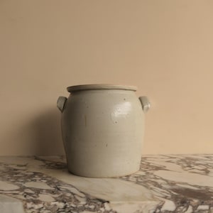 May include: A pale grey stoneware crock with two small handles. The cylindrical pot has a slightly flared rim and a smooth, aged appearance. The crock is likely used for food storage.